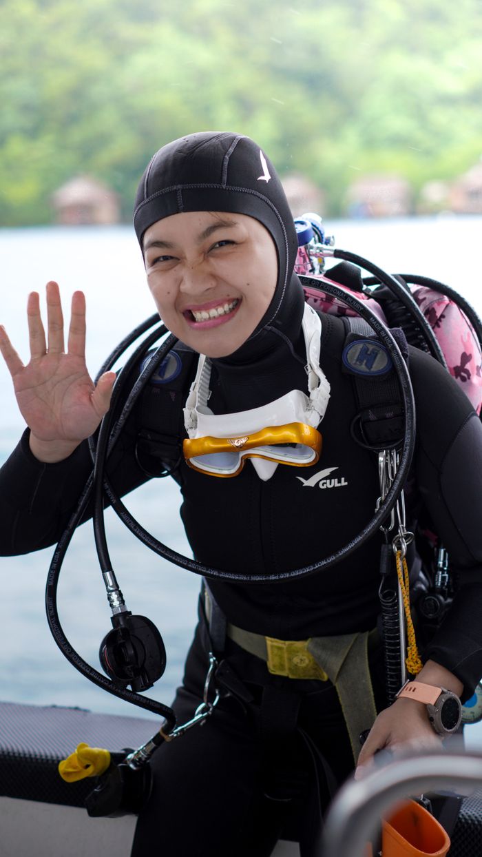 Smiling diver in wetsuit with gear waving on a boat, ready for a dive.